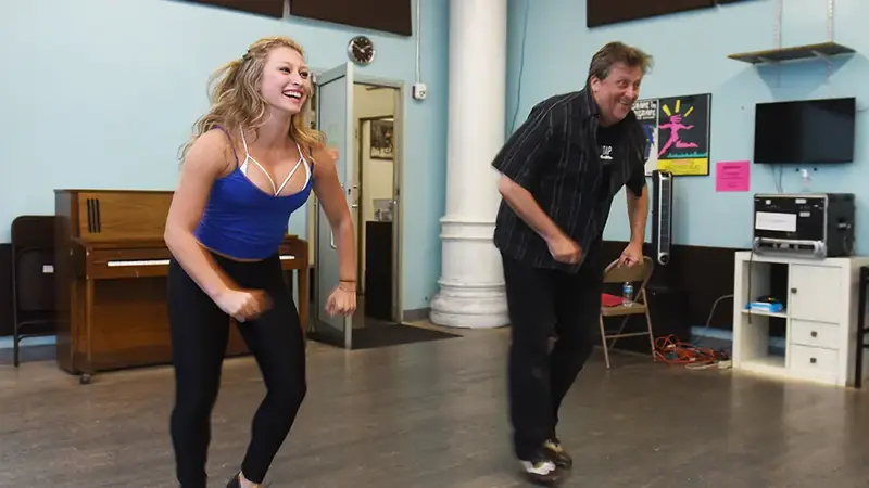 A young woman and an older man practice dance steps side by side in a bright studio with a piano and a wooden floor