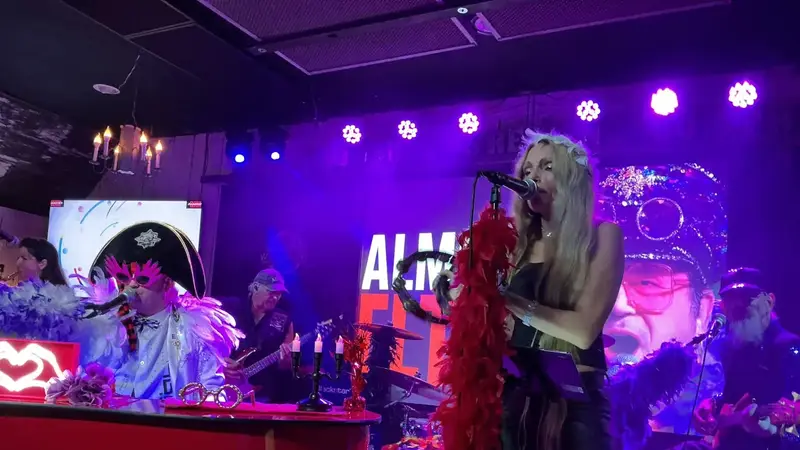 The tribute band on a club stage, with a singer in a red feather boa and the Elton performer in a sparkly cap at the piano