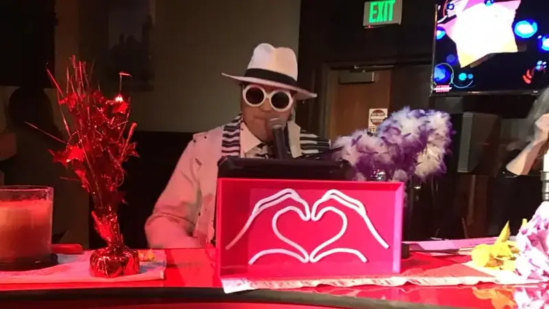 The Elton performer in a white hat and big white glasses singing at a red piano