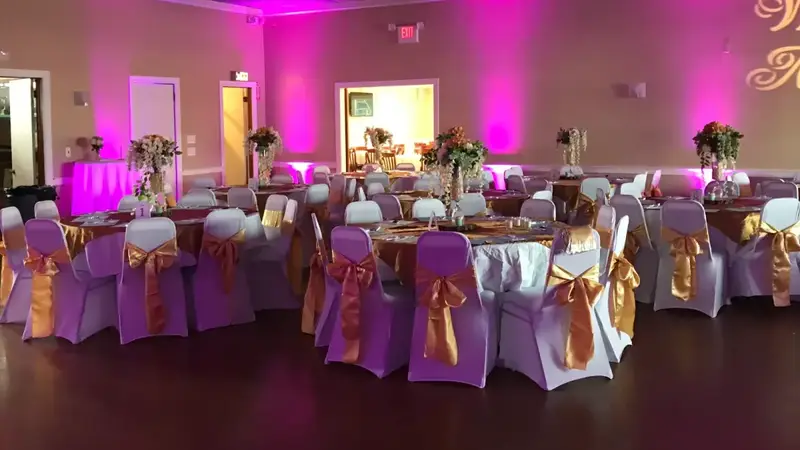 The main hall set for a party with round tables, white chair covers with gold bows and pink uplighting on the walls