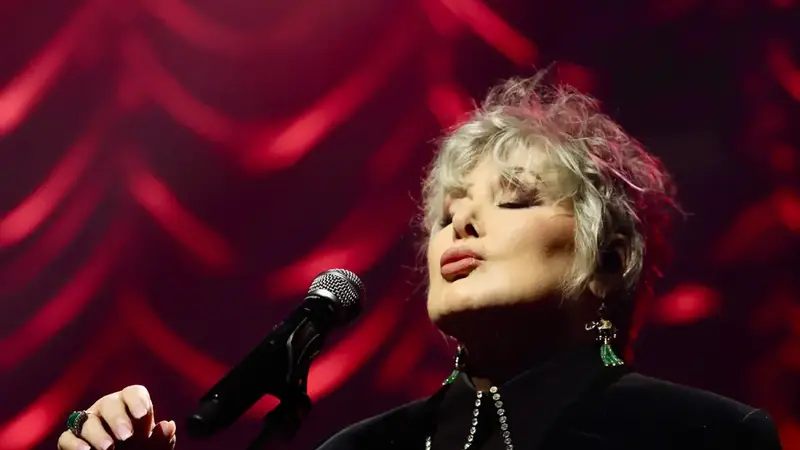 Ann Wilson singing into a microphone on a stand in front of a red curtain