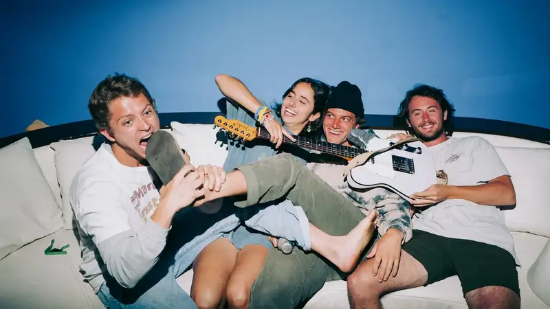 The four members of Arcy Drive goofing around on a white couch against a blue wall, one holding an electric guitar