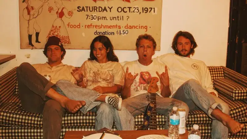 The four members of Arcy Drive sitting barefoot on a checkered couch under an old dance-night poster, one holding a bass