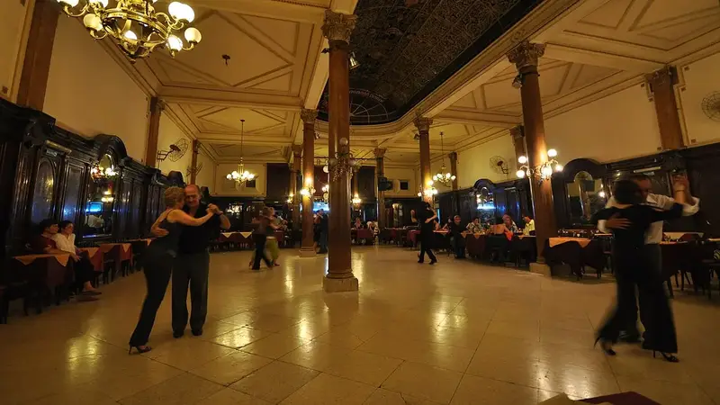 Couples dance tango on the marble floor of an old Buenos Aires ballroom with columns and chandeliers