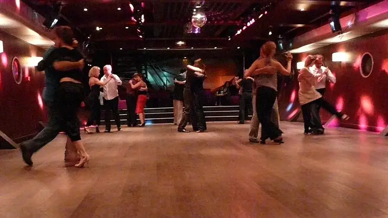 Couples in close embrace dance tango across a wide wooden floor in a dim club with pink lights