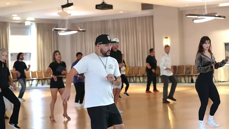 An instructor in a cap leads a group bachata class through a footwork pattern in a hotel ballroom