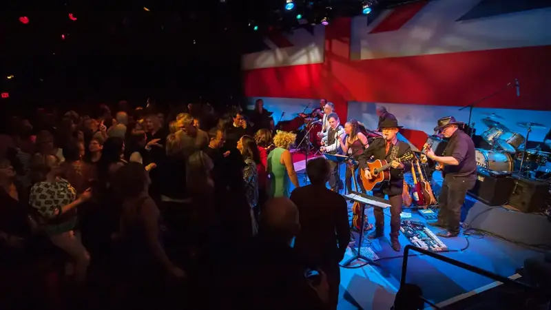 A band plays guitars on the Bay Street Theater stage in front of a British flag backdrop while a full house watches