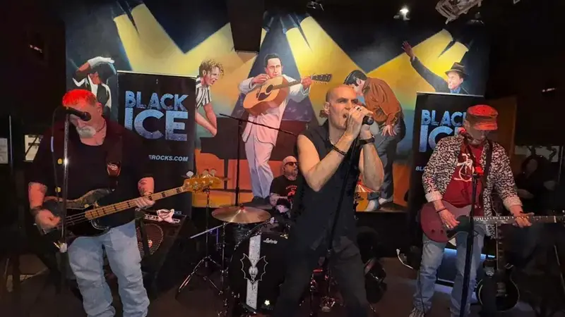 Black Ice playing in a bar: the singer at the mic between a bass player and a guitarist, with Black Ice banners and a mural behind