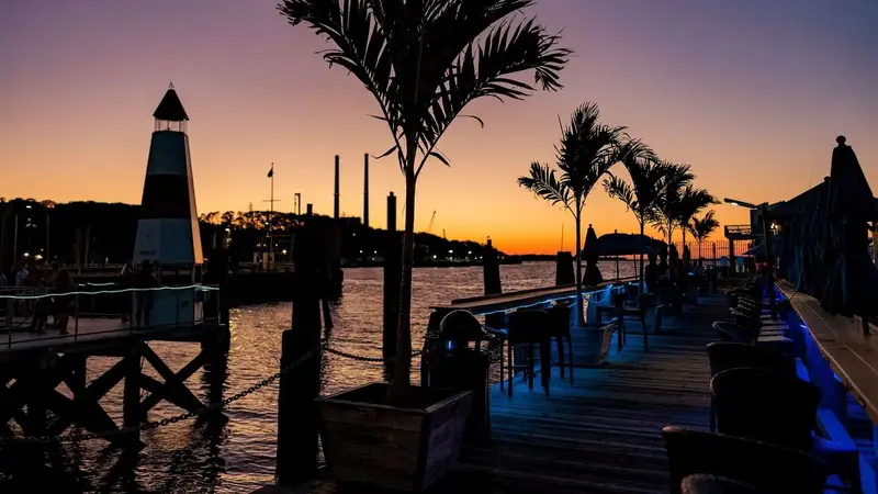 The waterfront dock deck at sunset, with potted palm trees, high-top tables lit in blue and a small lighthouse on the harbor