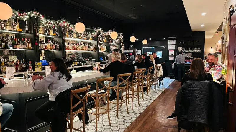 Diners at a long bar with wood stools, a lit wall of bottles and globe lights over a tiled floor