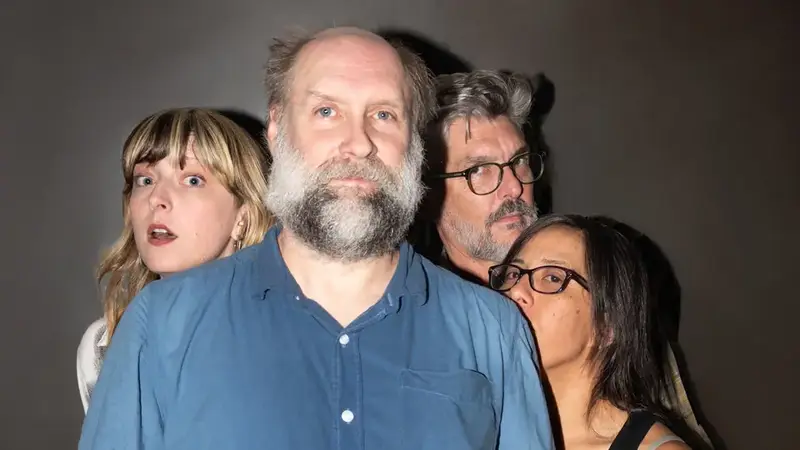 Four members of Built to Spill crowded close together against a gray wall, a bearded man in a blue shirt in front