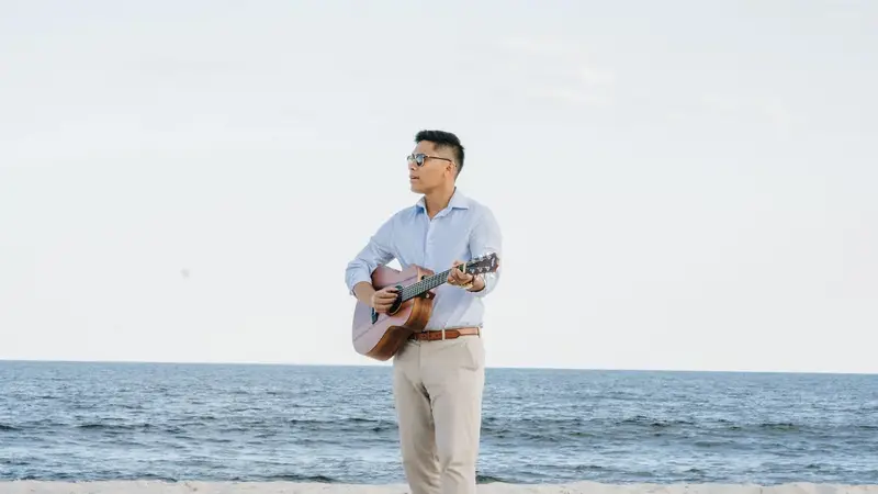 Bandleader Christian Montanez standing barefoot on a beach holding an acoustic guitar