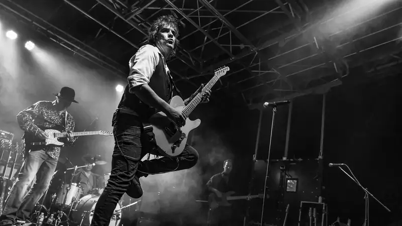 Black-and-white photo of the Classic Stones Live guitarist kicking up a leg while playing, with the band behind him