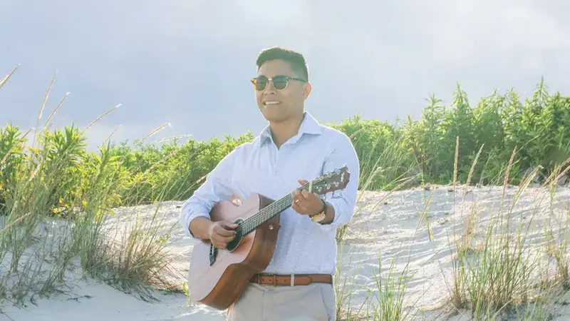 Christian Montanez in sunglasses, barefoot on a sand dune, strumming an acoustic guitar