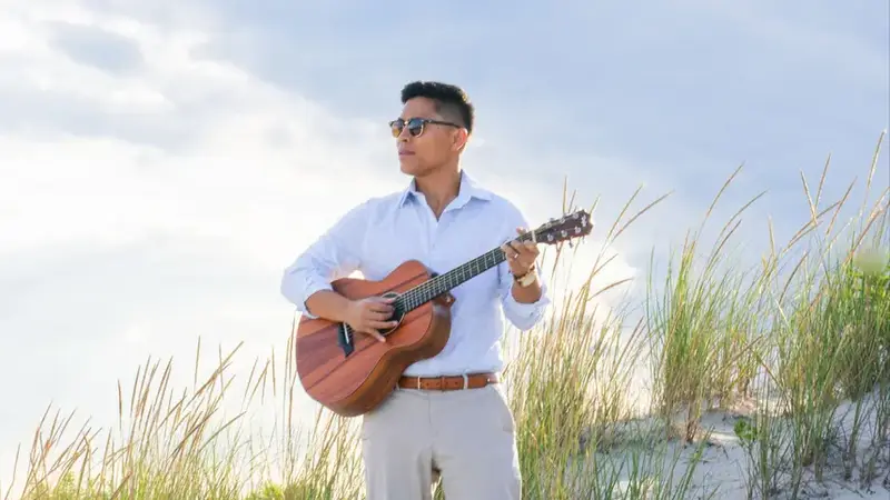 Christian Montanez playing an acoustic guitar among beach grass on a sand dune under a cloudy sky