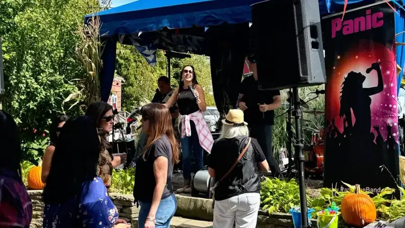 A cover band playing under a blue tent on the lawn at the Cooperage Inn Fall Festival, with guests standing in front and pennant flags overhead