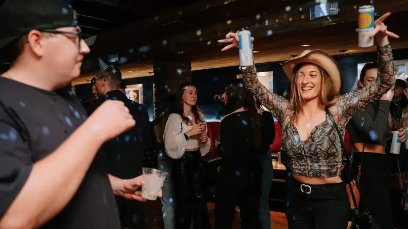 A woman in a cowboy hat dances with her arms up among a crowd on a wood dance floor with red booths behind