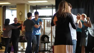 Adult students in pairs practicing a partner dance during a group class on a wooden studio floor