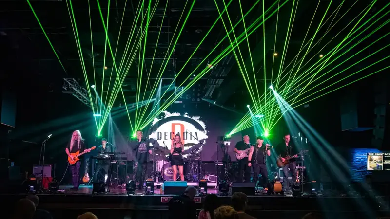 Decadia playing on a big club stage under fans of green laser beams, with the band's logo on the screen behind them