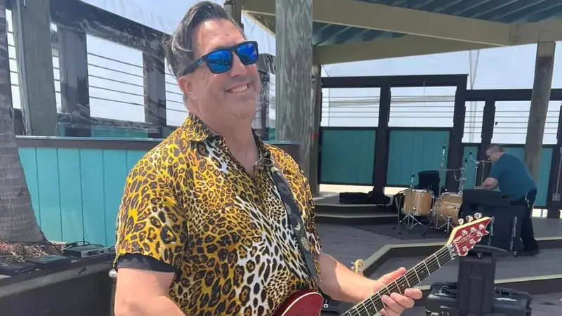 Dock Holiday guitarist in a leopard-print shirt and sunglasses playing a red guitar on a sunny waterfront deck