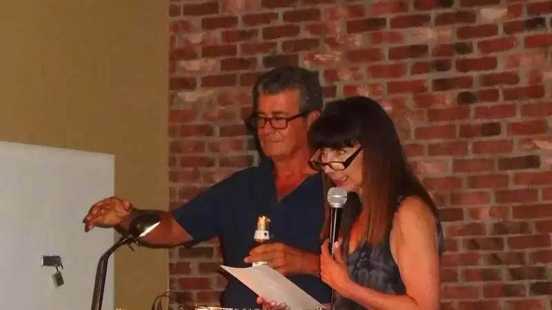 Donna DeSimone speaks into a microphone at a DJ table on stage, next to a fellow DJ, in front of a brick wall