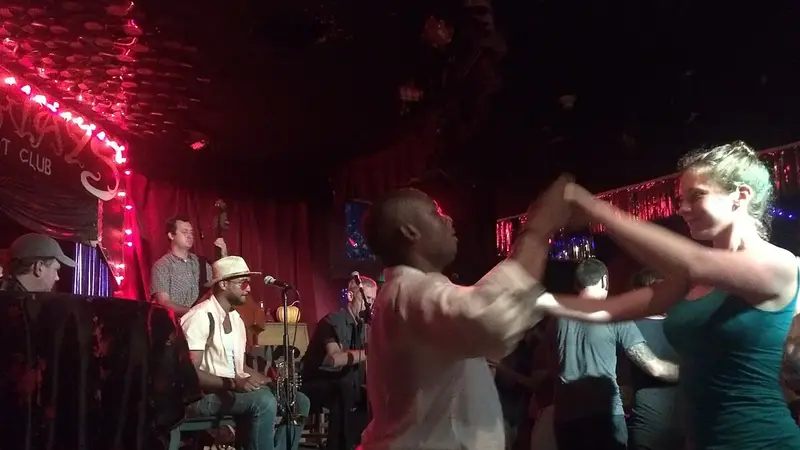 Couples swing dance in a dim, red-lit lounge while a small jazz band plays on stage