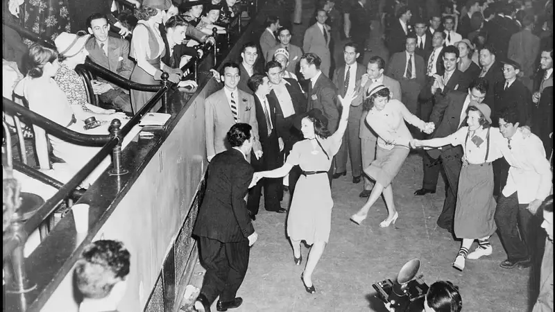 Black-and-white photo of couples jitterbugging on a crowded New York dance floor as onlookers watch and a photographer films