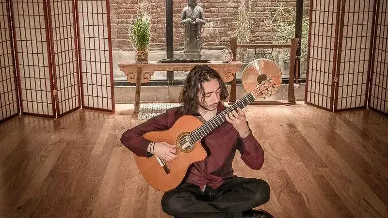 Eran Polat sits cross-legged on a wooden floor playing a classical guitar in a room with paper screens and a brick wall