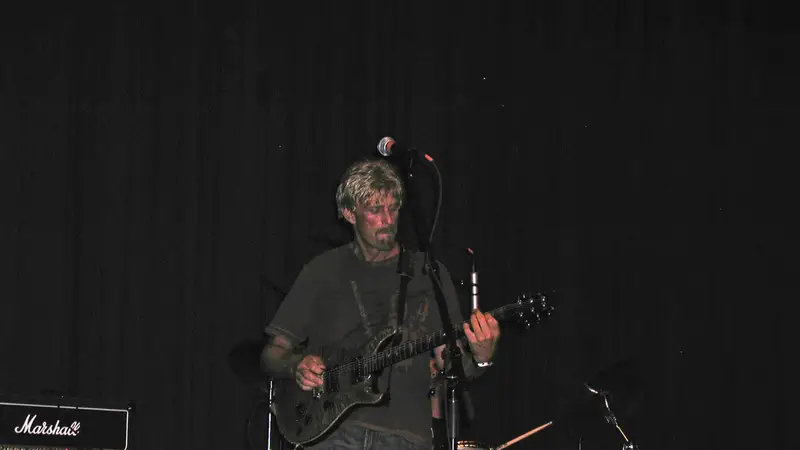 Erich Glaubitz singing into a microphone and playing an electric guitar on a dark stage, with a drum kit and Marshall amp behind him
