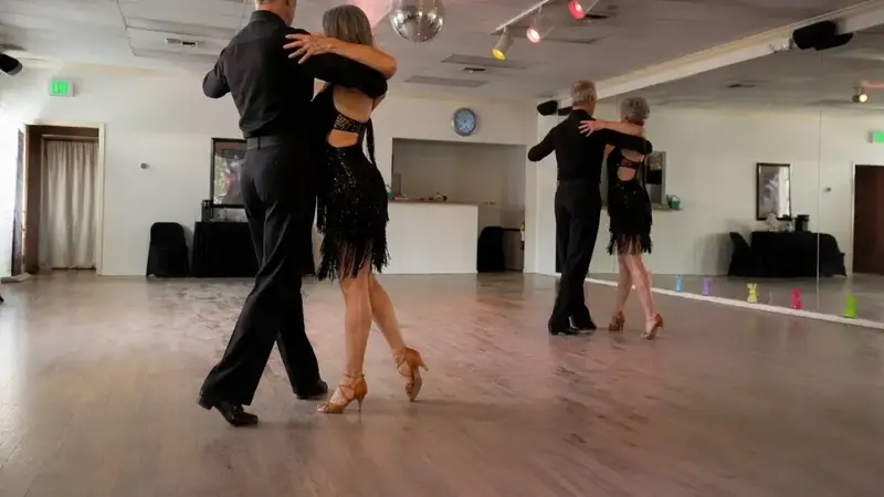 Two adult couples in black dancewear dancing a Latin dance on the studio's wood floor
