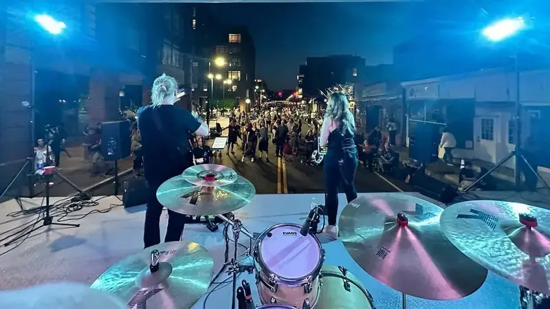 View from behind the drum kit of FiveStone playing an outdoor street-fair stage at night, with a crowd filling the street