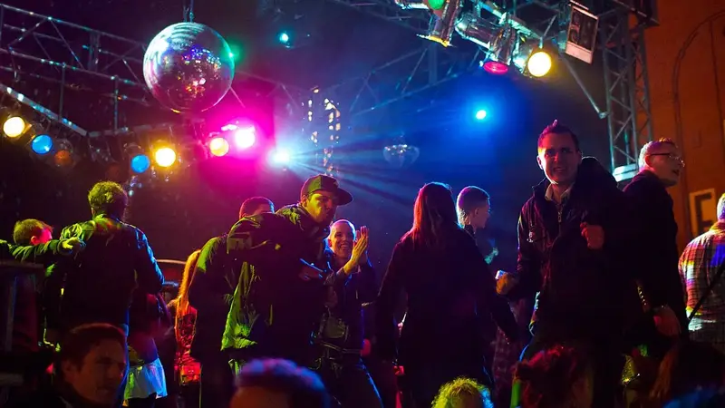 A crowd dances under a mirror ball and colorful stage lights at an open-air disco party