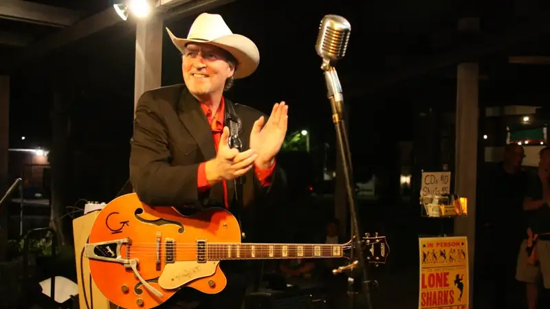 Gene Casey on stage at night with an orange guitar, clapping beside a vintage microphone