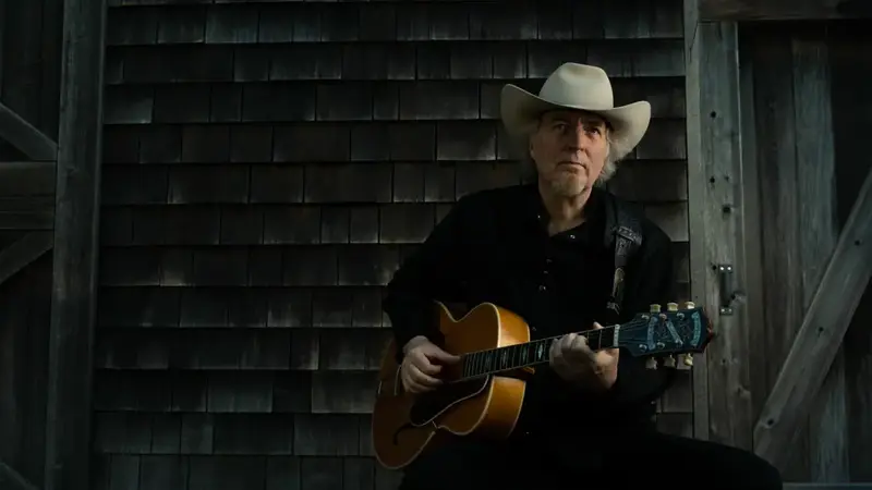 Gene Casey in a cowboy hat sitting with an acoustic guitar in front of a weathered shingled barn