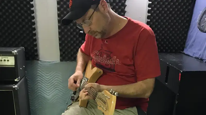 A band member in a red T-shirt and baseball cap playing electric guitar on a stool in the rehearsal studio