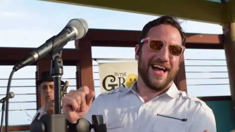 An In The Groove singer in sunglasses and a white shirt dancing at the microphone on an outdoor waterfront stage, with the band's banner behind him