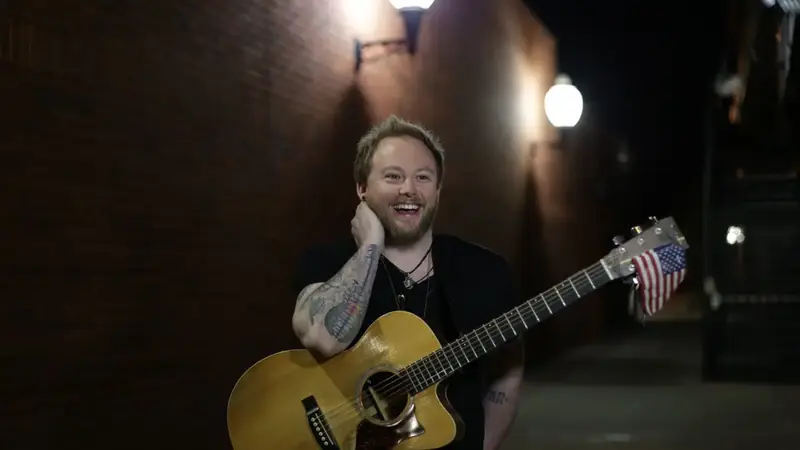 JD Leonard laughing on a brick-lined street at night, holding an acoustic guitar with a small American flag on the headstock