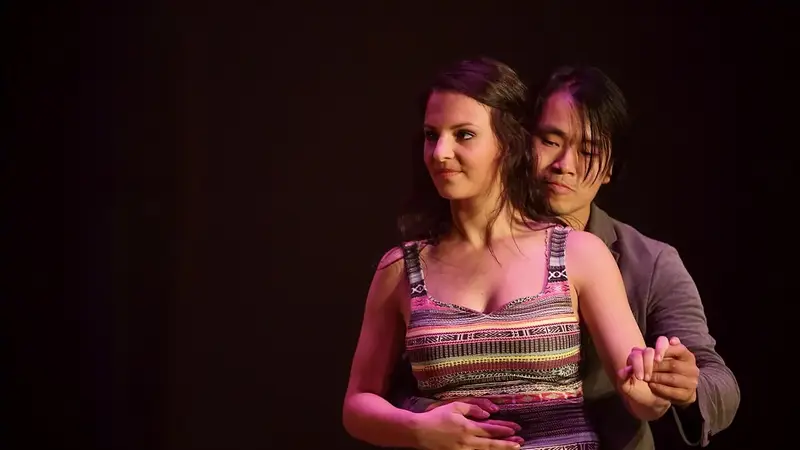 A dance couple performs a slow kizomba move on a dark stage, the man standing close behind the woman