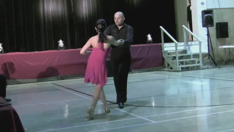 A man in black and a woman in a pink dress dance the rumba in a closed hold in a school hall