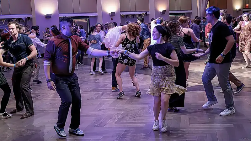 Couples swing out across a wooden ballroom floor; in front, a man in a red shirt and tie leads his smiling partner by the hand