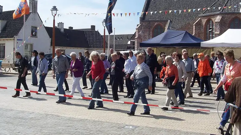 Older adults line dance in rows on a town square with flags and bunting, next to a brick church