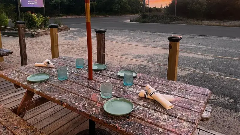 An outdoor picnic table set with plates and glasses outside Little Fish, under a pink and orange sunset sky
