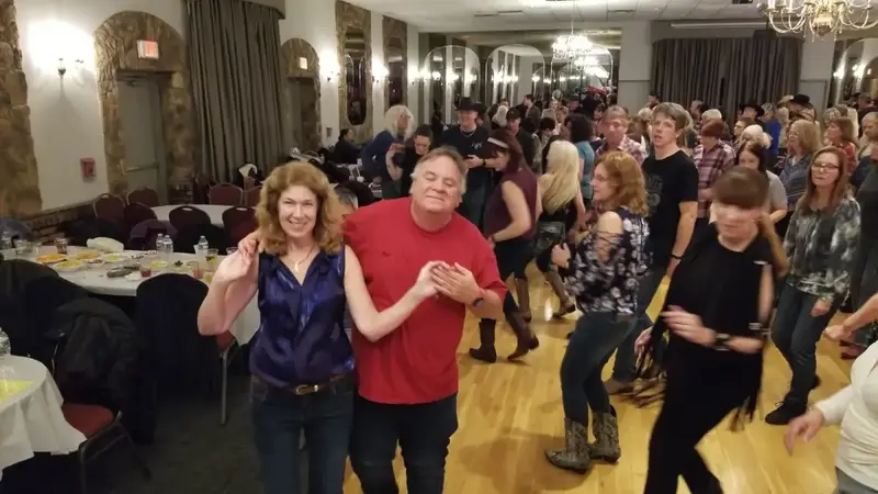 A crowded dance floor of smiling adults dancing under chandeliers in a banquet hall