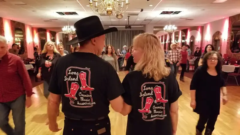 A couple in black LICMA T-shirts, one in a cowboy hat, walking hand in hand across the dance floor