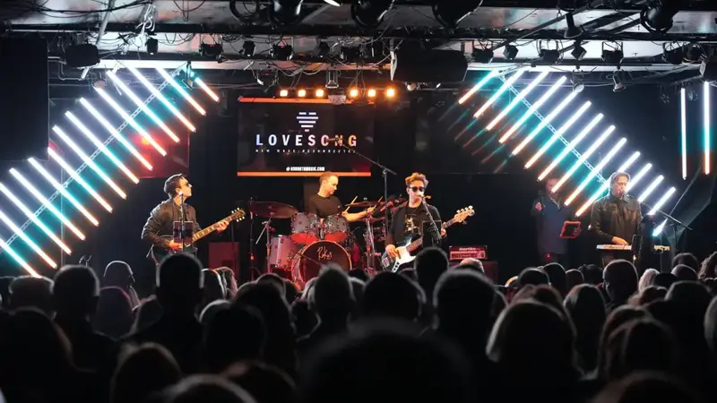 The full Lovesong band playing under a lit Lovesong sign, framed by slanted white light bars, seen over the heads of the audience