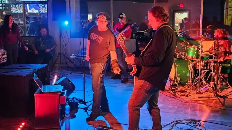 A rock band with a singer, guitarist, bassist and drummer playing on the lounge's wood floor under colored lights