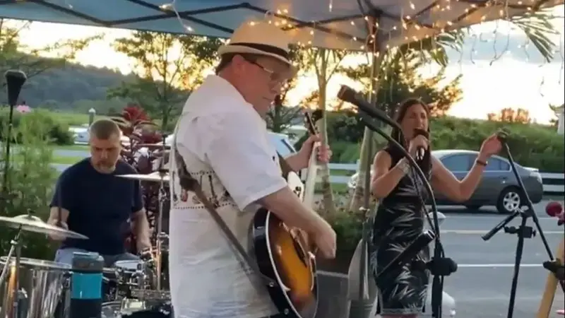 Mach5 performing outdoors under a canopy at sunset, a guitarist and a woman singing with a drummer behind them