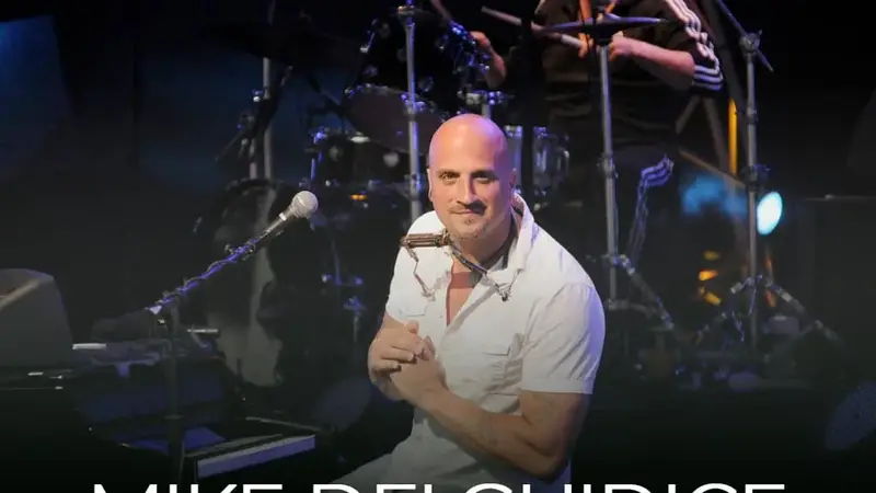 Mike DelGuidice in a white shirt smiling on stage in front of a drum kit under stage lights