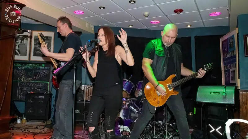Miss Mayhem's singer and two guitarists rocking out in a bar with colored lights on the floor