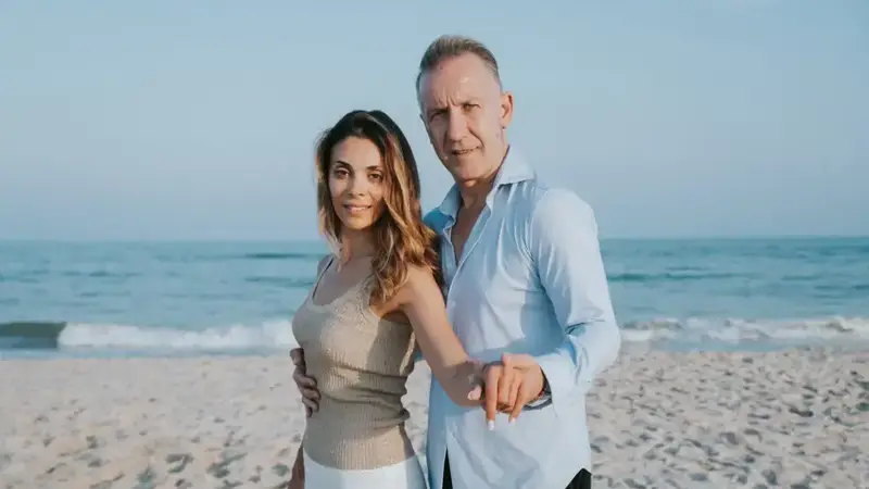 Monito holds hands with a dance partner in a ballroom pose on a sandy beach with the ocean behind them